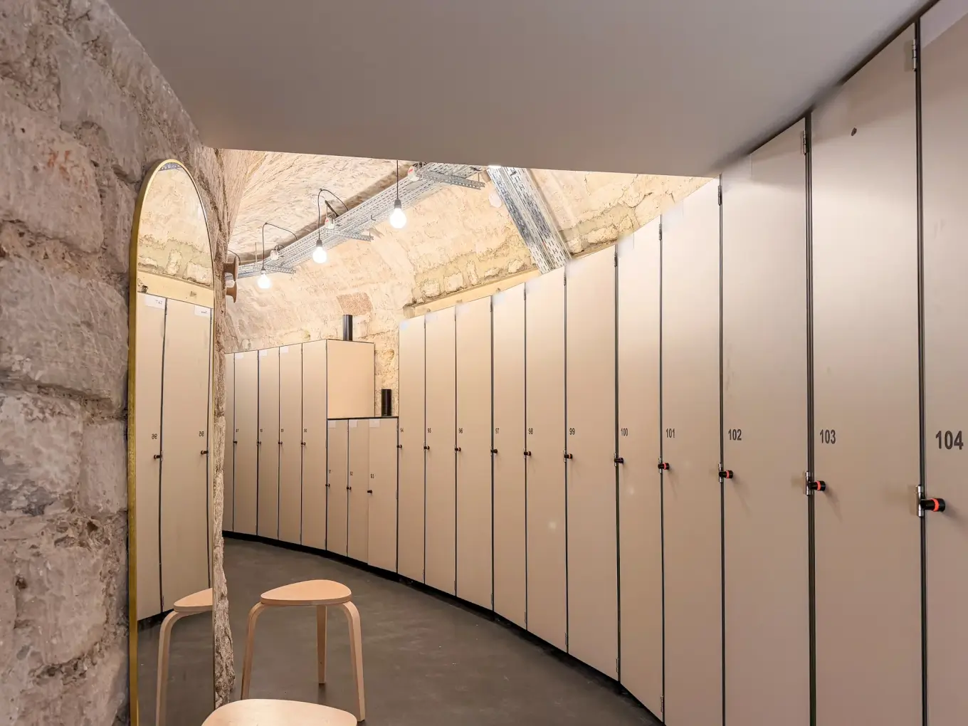 Rangée courbe de casiers de change à une porte, numérotés, dans une galerie voûtée en pierre ; miroir et tabourets au premier plan