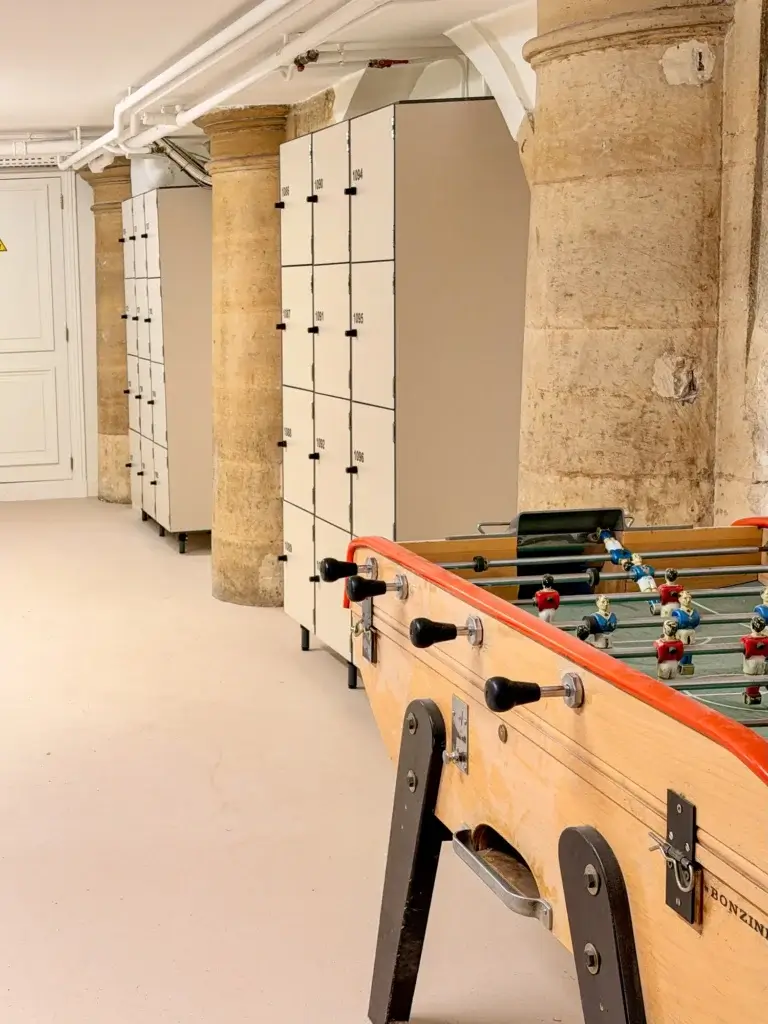 Deux blocs de casiers de rangement à douze cases, sur pieds, installés entre des piliers en pierre ; un baby-foot au premier plan