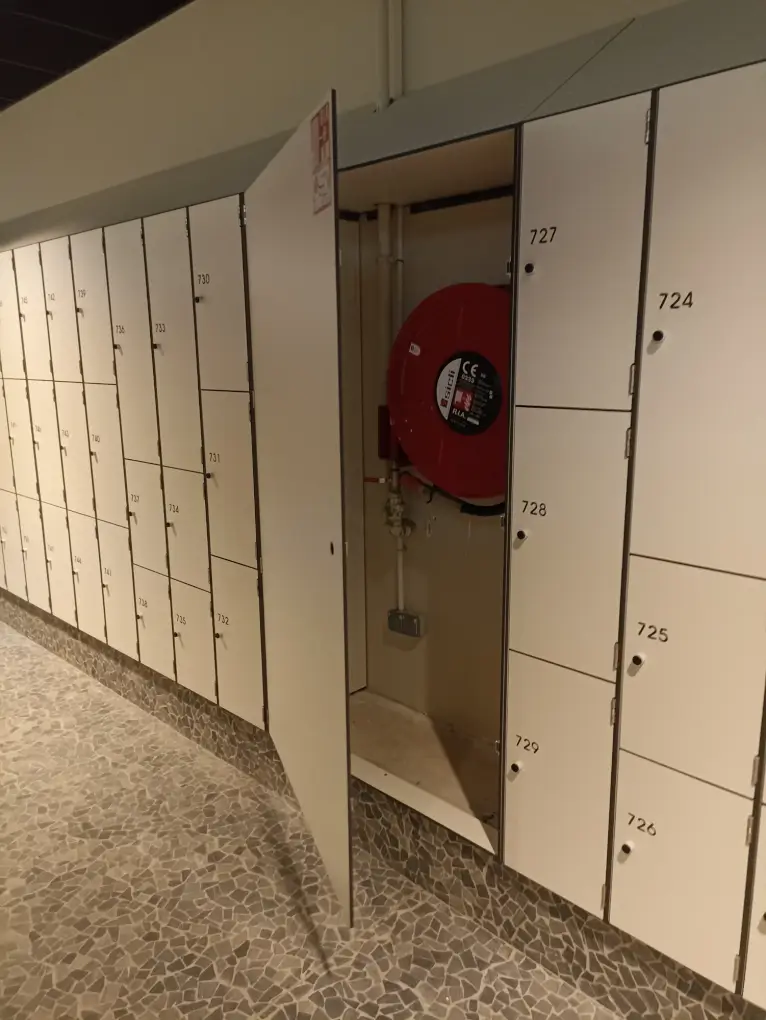 Casiers blancs numérotés sous un toit incliné, avec une porte toute hauteur ouverte sur un robinet d'incendie