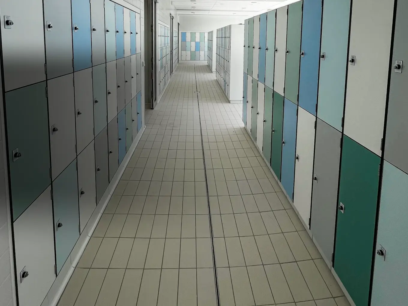 Allée de vestiaire du centre aquatique de Valognes : casiers encastrés dans le mur à gauche, îlot de casiers à droite, portes blanches, grises, bleues et vertes